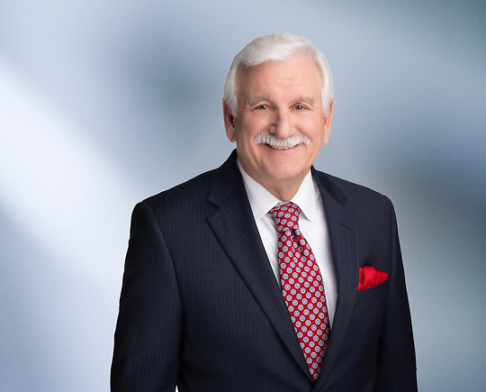 A man, Michael Kluck, with short, graying hair, wearing a dark suit and striped tie, is smiling at the camera, standing against a blurred blue and white background."